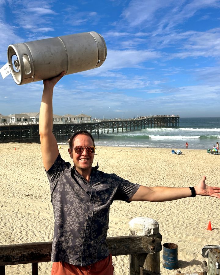 A Perpetual founder holding a Steady State Roasting cold brew keg overhead on the beach beside Crystal Pier in Pacific Beach